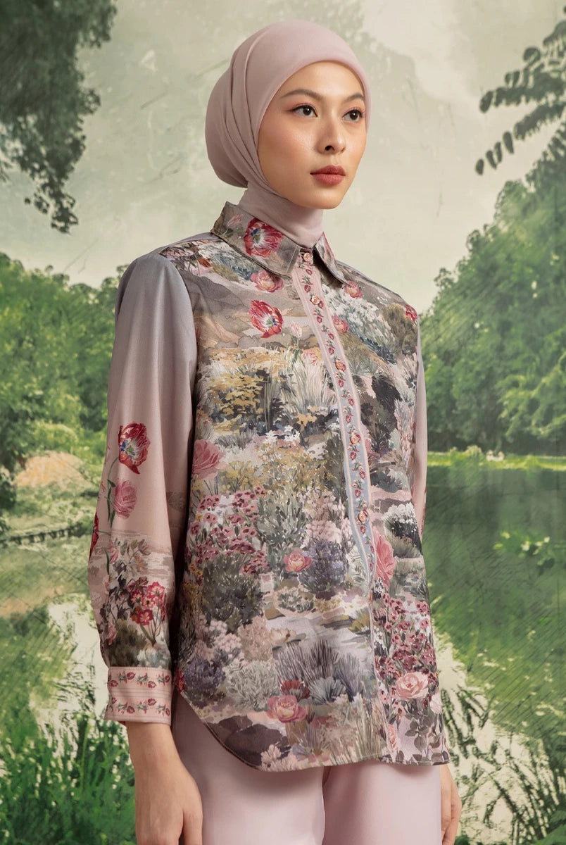 A woman wearing a light pink hijab and a matching long-sleeve button-up shirt by Hijabchic with a detailed multicolor landscape print on the front and sleeves, set against a scenic forest background.
