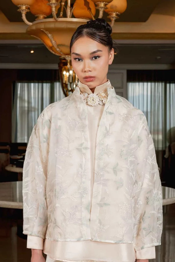 A woman stands indoors wearing an ash grey-colored embroidered jacket by Jenna & Kaia with a high collar and a decorative floral applique at the neck, paired with a matching cream satin garment. The setting is a luxurious interior with warm lighting, chandeliers, and elegant furniture.