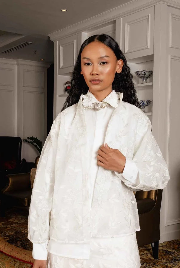 A woman stands indoors wearing a broken white-colored embroidered jacket by Jenna & Kaia with a high collar and a decorative floral applique at the neck, paired with a matching cream satin garment. The setting is a luxurious interior with warm lighting, chandeliers, and elegant furniture.