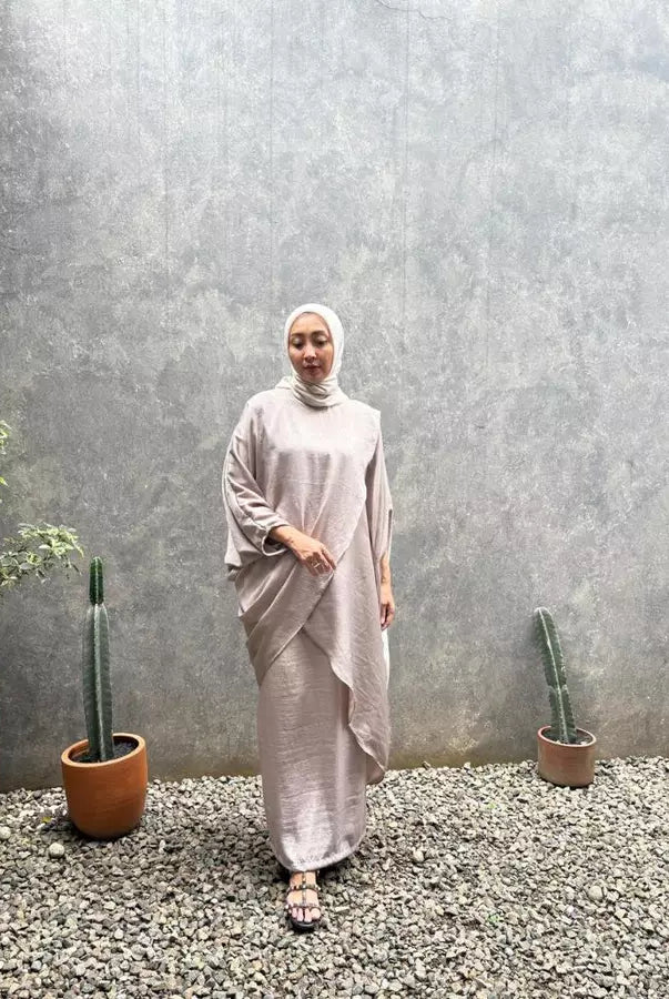 Woman in a light-colored outfit set by Abame standing in front of a textured wall with potted plants.