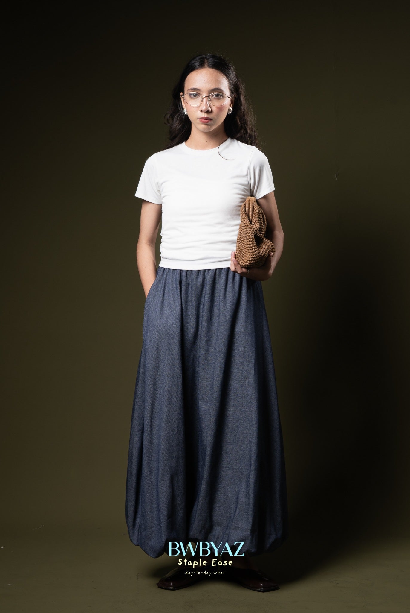 Woman wearing a white t-shirt and long blue skirt from BWBYAZ holding a brown bag against a dark background