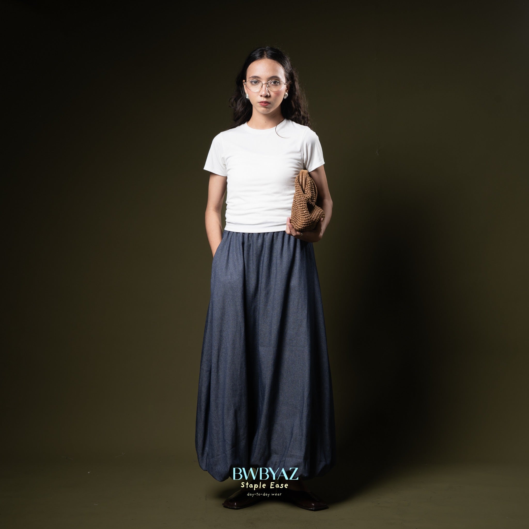 Woman wearing a white t-shirt and long blue skirt from BWBYAZ holding a brown bag against a dark background