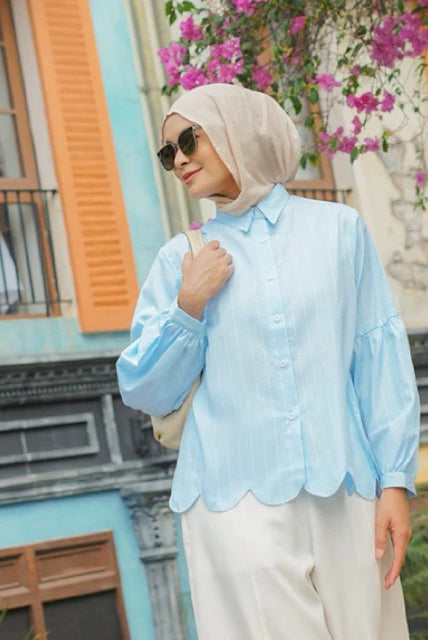 Woman wearing a light blue shirt by Casa Elana and beige hijab in front of a colorful building with flowers.