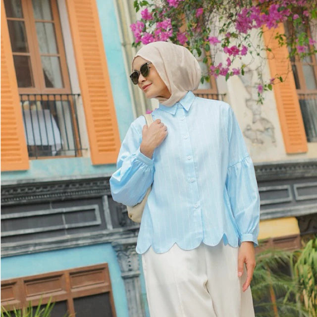 Woman wearing a light blue shirt by Casa Elana and beige hijab in front of a colorful building with flowers.