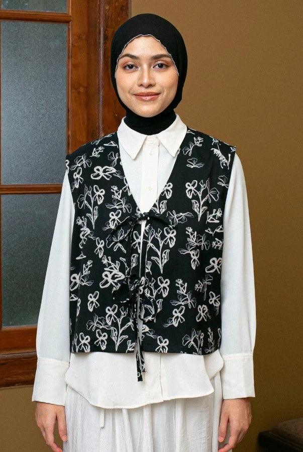 Woman wearing a black floral vest by Lozy Hijab over a white shirt in front of a window.