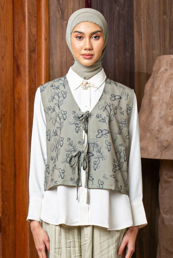 Woman wearing a patterned sage vest by Lozy Hijab over a white shirt in front of a wooden door.