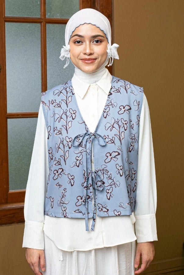 Woman wearing a floral sky blue vest by Lozy Hijab over a white shirt in a room with a wooden door.