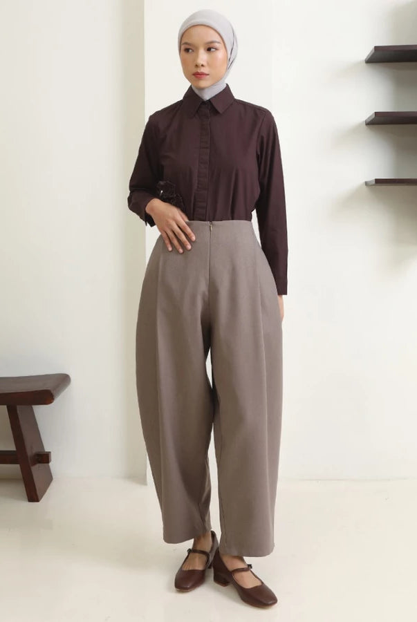 Woman wearing a brown shirt and mocca wide-leg pants by Hijabchic standing in a minimalistic room.