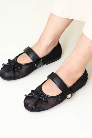 A pair of women's flat shoes by Dianable with lace detailing and small bows on the top, color black, worn by a person standing.