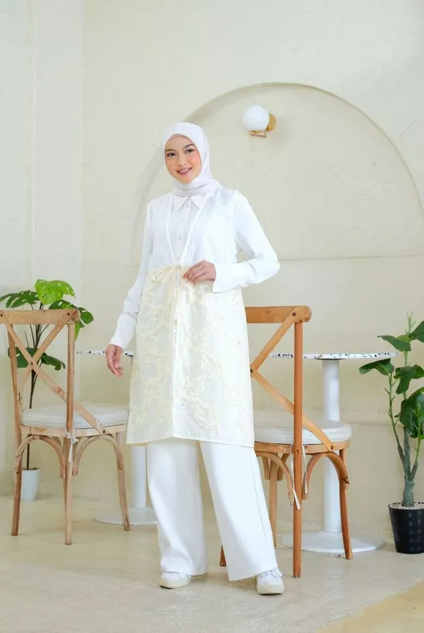 Woman wearing a white vest tunic by Abame standing in a minimalistic room with plants and chairs.