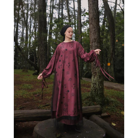 A woman wearing a long, printed dress by Maima with long sleeves in a maroon color, standing on a pathway in a forested area.