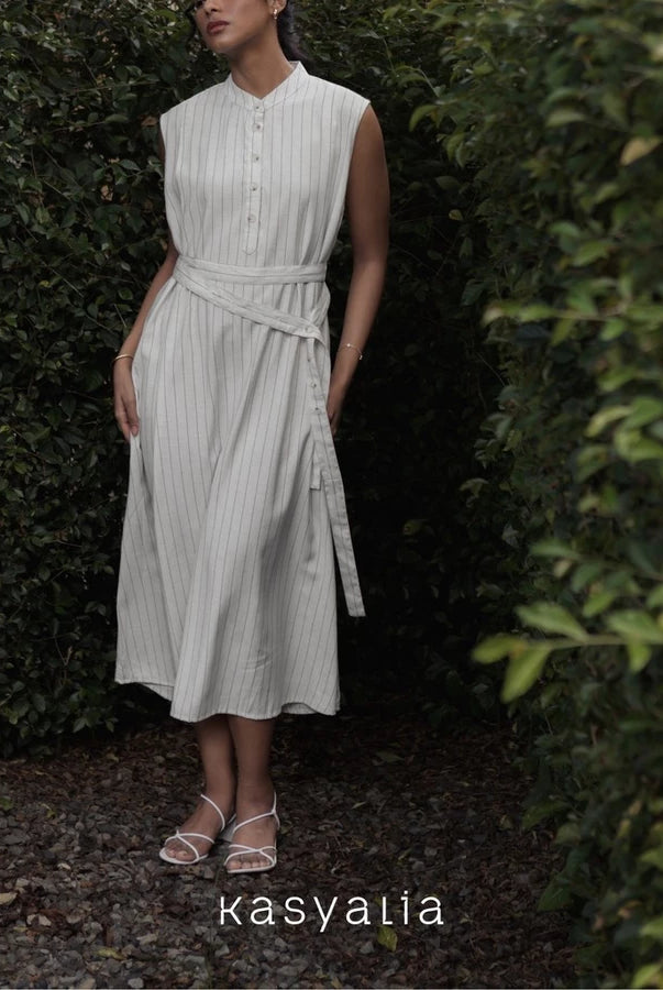 Woman wearing a light-colored dress with a belt standing in a garden setting, with 'kasyalia' branding.