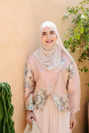 Woman wearing a pink floral top by Haidee Orlin and hijab standing in front of a beige wall with plants.