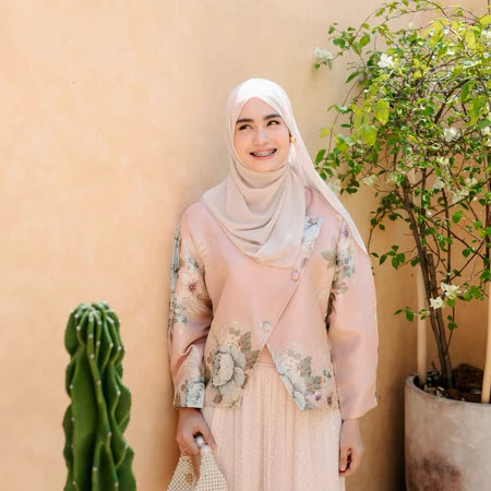 Woman wearing a pink floral top by Haidee Orlin and hijab standing in front of a beige wall with plants.