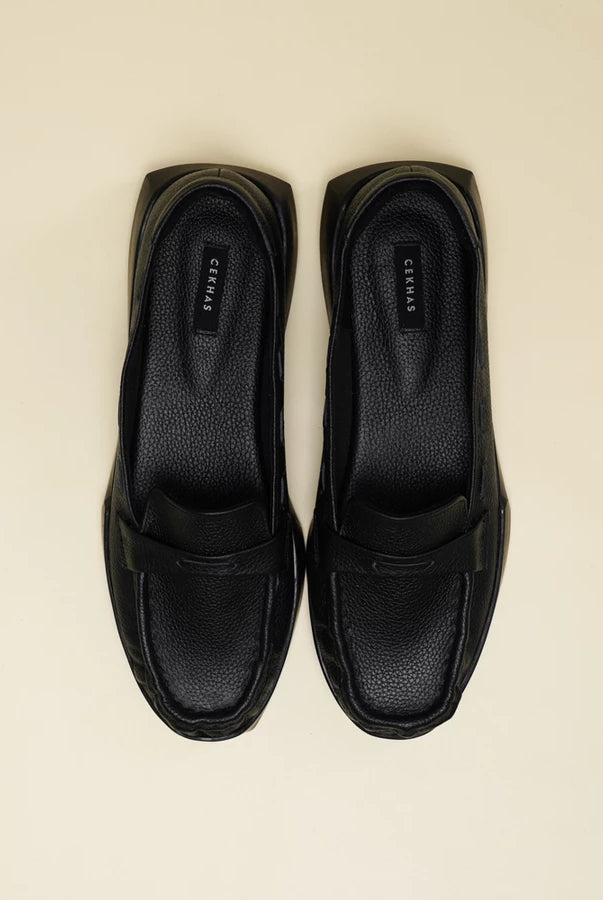 A pair of black leather penny loafers flatshoes with a chunky black sole, photographed from a top-down view on a light beige background. The brand name 'Cekhas' is visible on the insole.