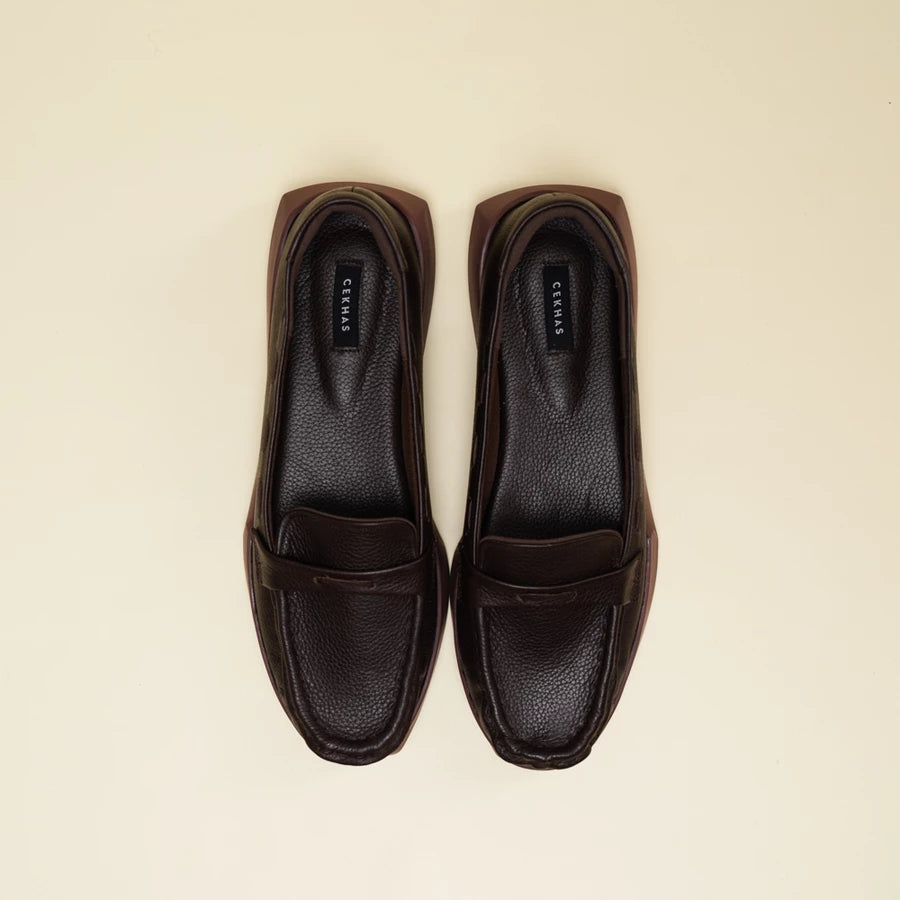 A pair of dark brown leather penny loafers flatshoes with a pebbled texture, shown from a top-down view on a light cream background. The brand name 'Cekhas' is visible on the insoles.