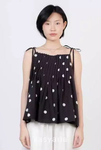 A model wearing a black sleeveless top by Kasyalia with white polka dots and white pants, standing against a plain light gray background.