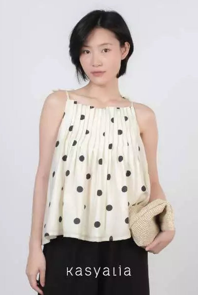 A woman modeling a cream-colored sleeveless top with black polka dots, paired with black pants and holding a woven straw clutch bag. The brand name 'kasyalia' is printed at the bottom.
