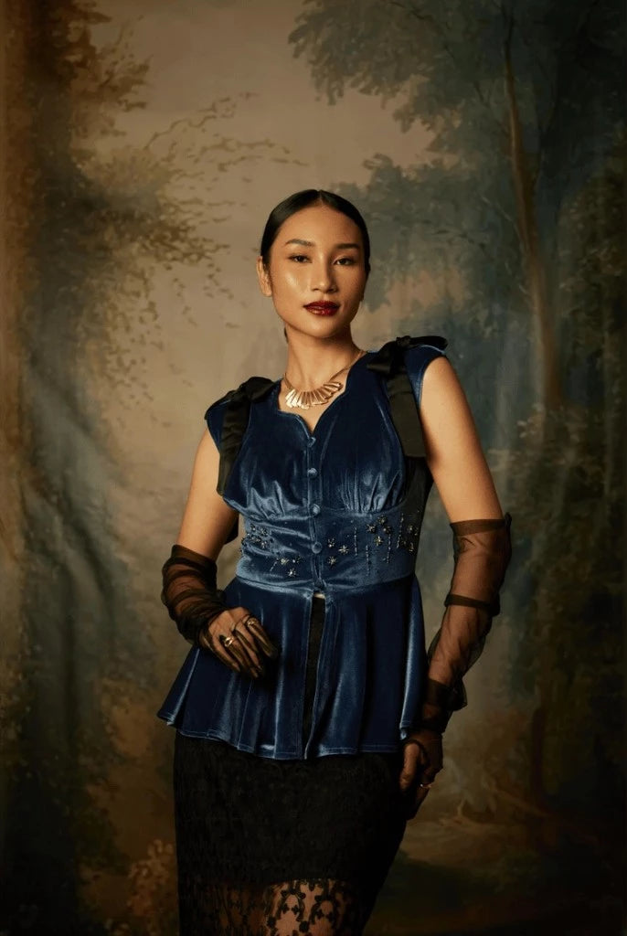 A woman wearing a sleeveless, flared, mid-night blue colored top by Kartinis Label with velvet material and decorative elements on the shoulders and waist.
