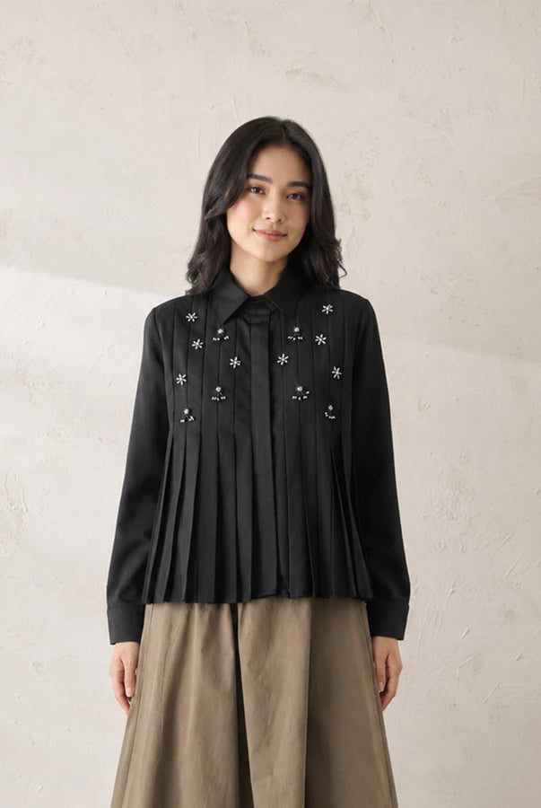 A woman stands against a textured beige wall wearing a black long-sleeve pleated shirt by Jenna & Kaia with embroidered white floral details and a bow at the collar, paired with a beige skirt.