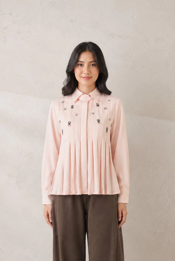 A woman stands against a textured light beige wall wearing a dusty pink pleated shirt by Jenna & Kaia with small dark embroidered details on the chest and dark brown wide-leg pants.