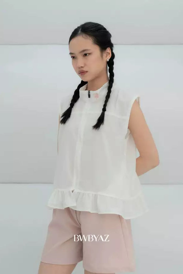 A young girl with braided hair wearing a white sleeveless vest by BWBYAZ with a ruffled hem and light pink shorts, standing against a light gray studio background. The brand name 'BWBYAZ' is printed at the bottom.