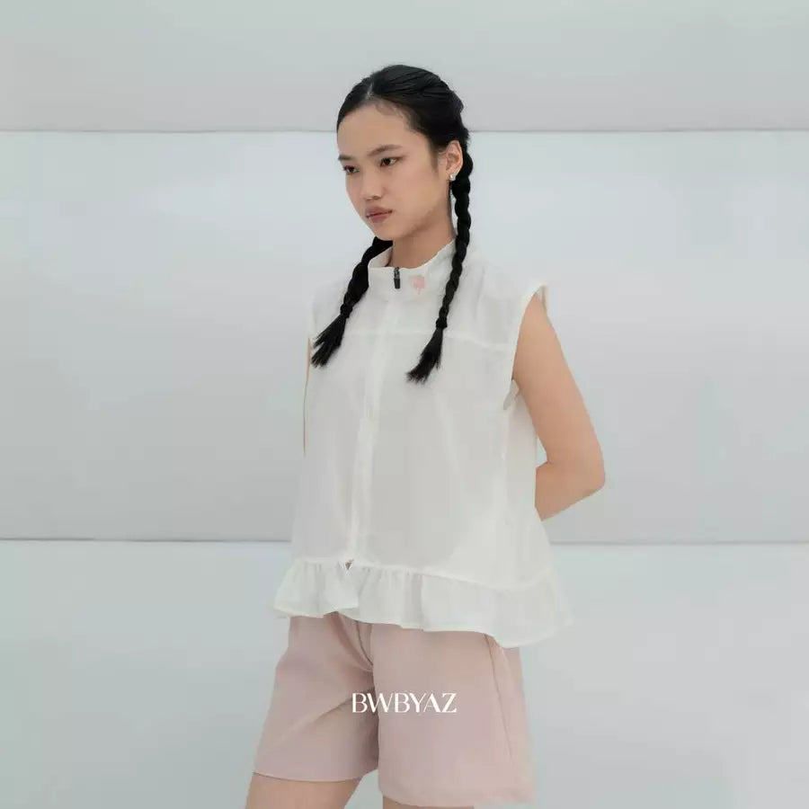 A young girl with braided hair wearing a white sleeveless vest by BWBYAZ with a ruffled hem and light pink shorts, standing against a light gray studio background. The brand name 'BWBYAZ' is printed at the bottom.