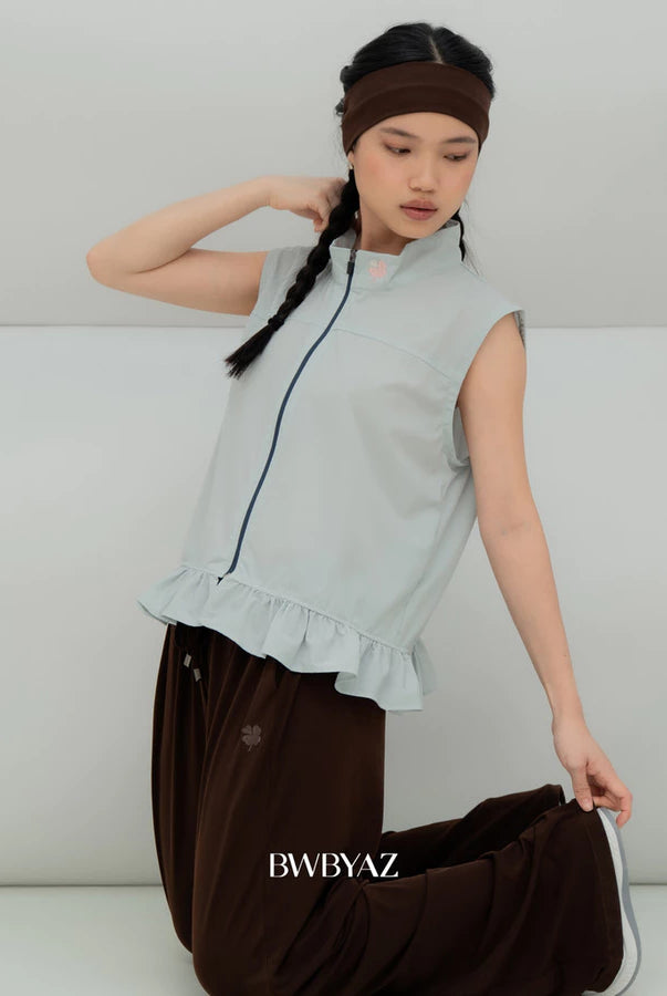 A model wearing a light aqua blue sleeveless vest by BWBYAZ with a ruffled hem and dark brown pants, kneeling against a minimalist light gray background. The brand name 'BWBYAZ' is printed on the pants.