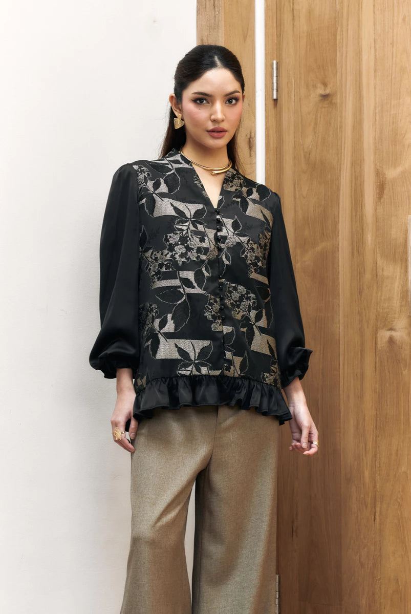 Woman wearing a black floral blouse by Studio Tui and beige pants against a wooden door.