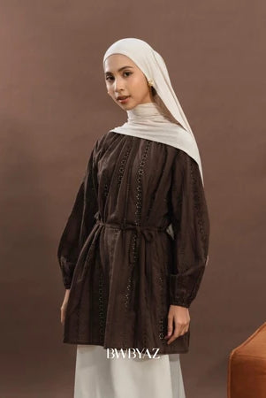 A woman wearing an elegant premium cotton lace tunic in brown from BWBYAZ, styled with a matching hijab and ivory skirt. The reversible tunic features delicate lace details, tie accents at the neckline, and bishop sleeves with elastic cuffs, paired with a small metallic gold handbag for a refined, versatile, and modest fashion look.