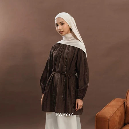 A woman wearing an elegant premium cotton lace tunic in brown from BWBYAZ, styled with a matching hijab and ivory skirt. The reversible tunic features delicate lace details, tie accents at the neckline, and bishop sleeves with elastic cuffs, paired with a small metallic gold handbag for a refined, versatile, and modest fashion look.