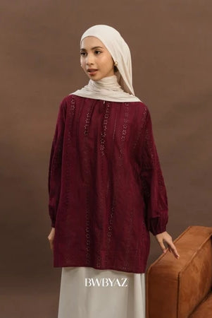 A woman wearing an elegant premium cotton lace tunic in maroon from BWBYAZ, styled with a matching hijab and ivory skirt. The reversible tunic features delicate lace details, tie accents at the neckline, and bishop sleeves with elastic cuffs, paired with a small metallic gold handbag for a refined, versatile, and modest fashion look.