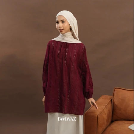 A woman wearing an elegant premium cotton lace tunic in maroon from BWBYAZ, styled with a matching hijab and ivory skirt. The reversible tunic features delicate lace details, tie accents at the neckline, and bishop sleeves with elastic cuffs, paired with a small metallic gold handbag for a refined, versatile, and modest fashion look.