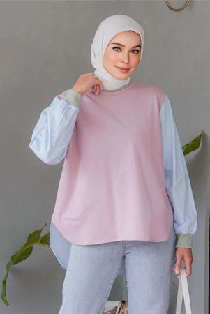 Woman wearing a light pink shirt with light blue sleeves by Casa Elana and light blue pants, standing indoors.