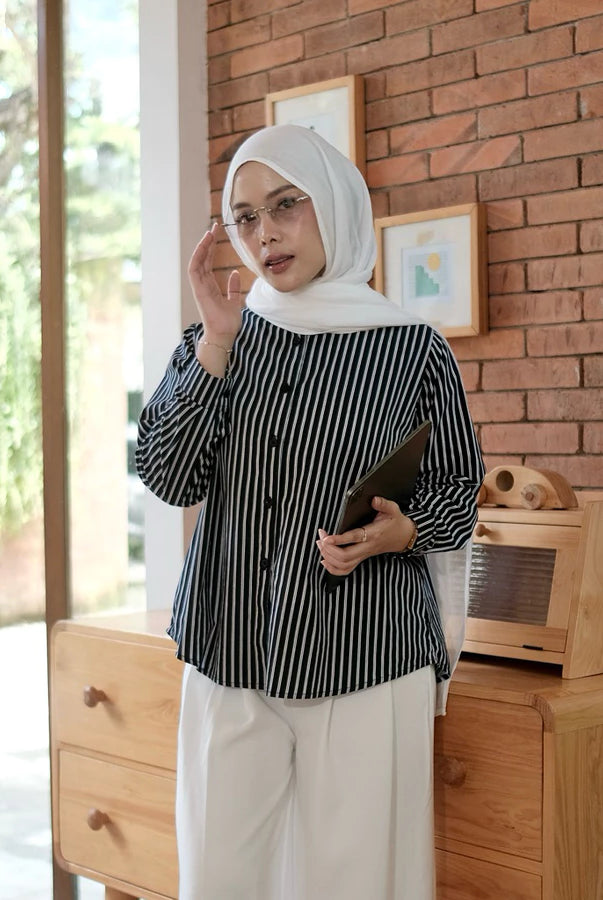 Woman wearing a black and white striped shirt by Renaette and hijab standing in a room with wooden furniture and a brick wall.