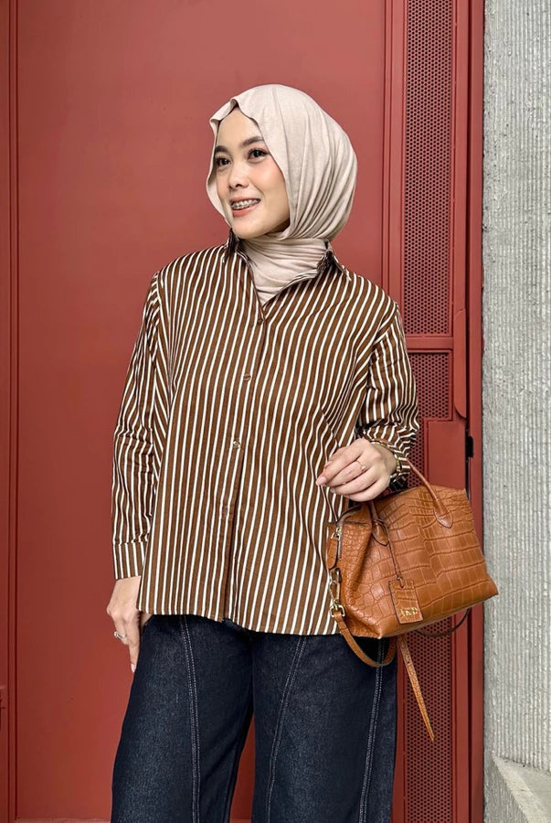 Woman wearing a brown striped shirt by Renaette and hijab, holding a brown leather bag against a red door.