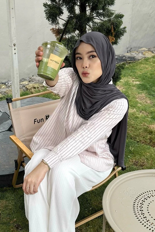 Woman in pink striped shirt by Renaette sitting outdoors holding a green cup, with a casual setting in the background.
