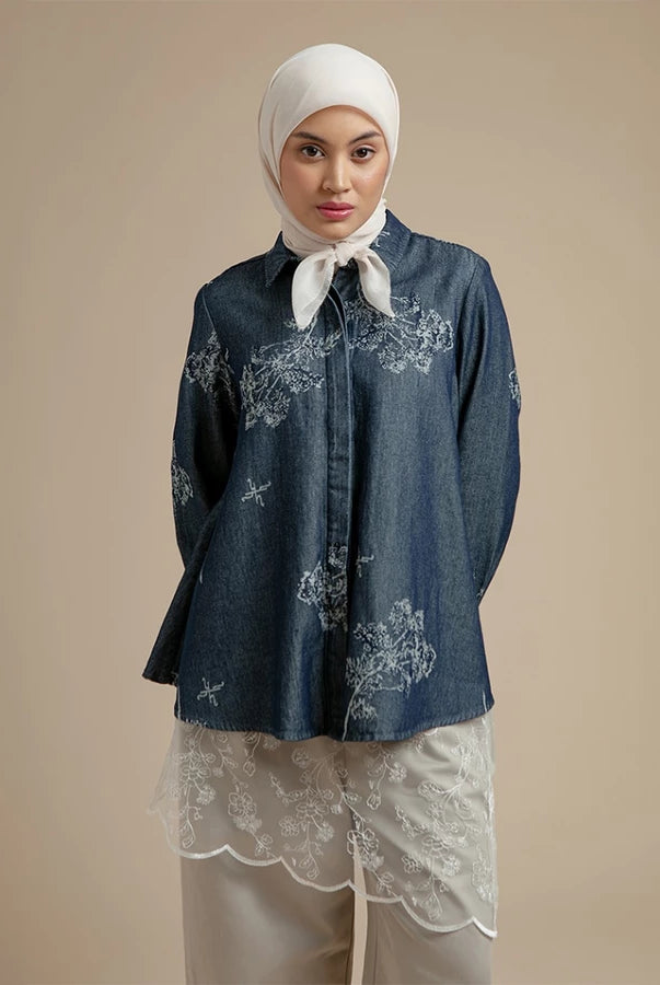 Woman wearing a dark denim shirt by Hijabchic with floral embroidery and a white hijab on a beige background