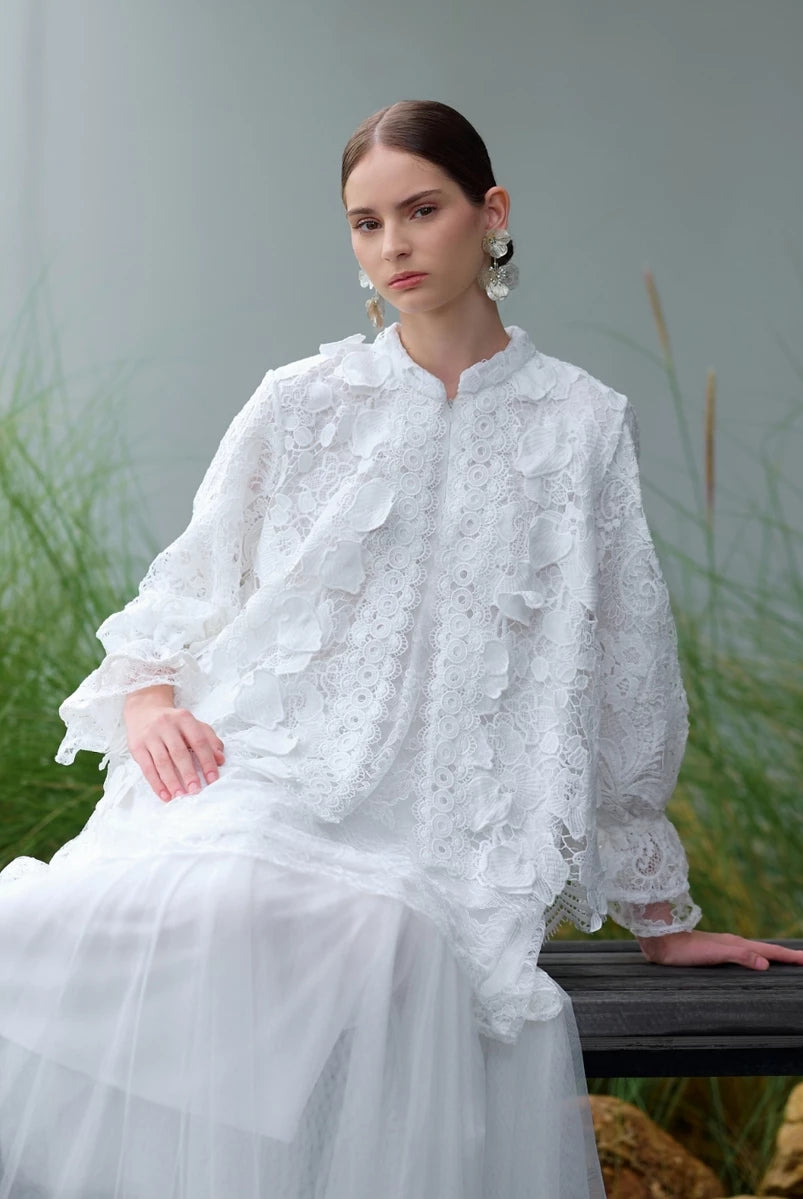 Woman in a white lace blouse by Kasa Heritage sitting outdoors with greenery in the background.