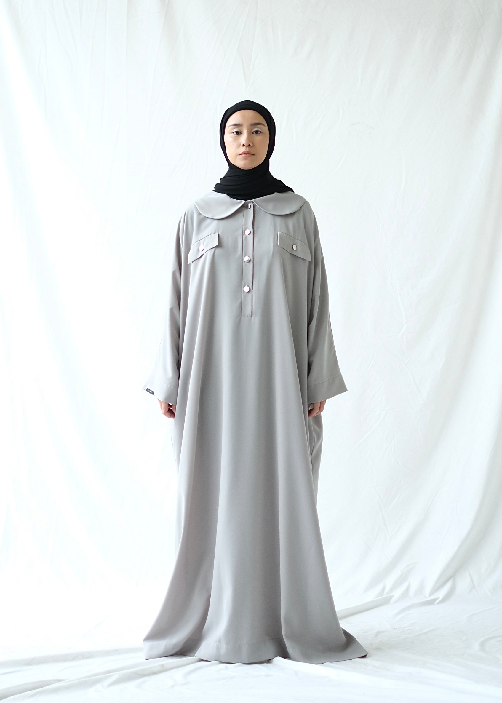 A person wearing a grey abaya by Rashawl with a button opening on the front, standing against a white background.