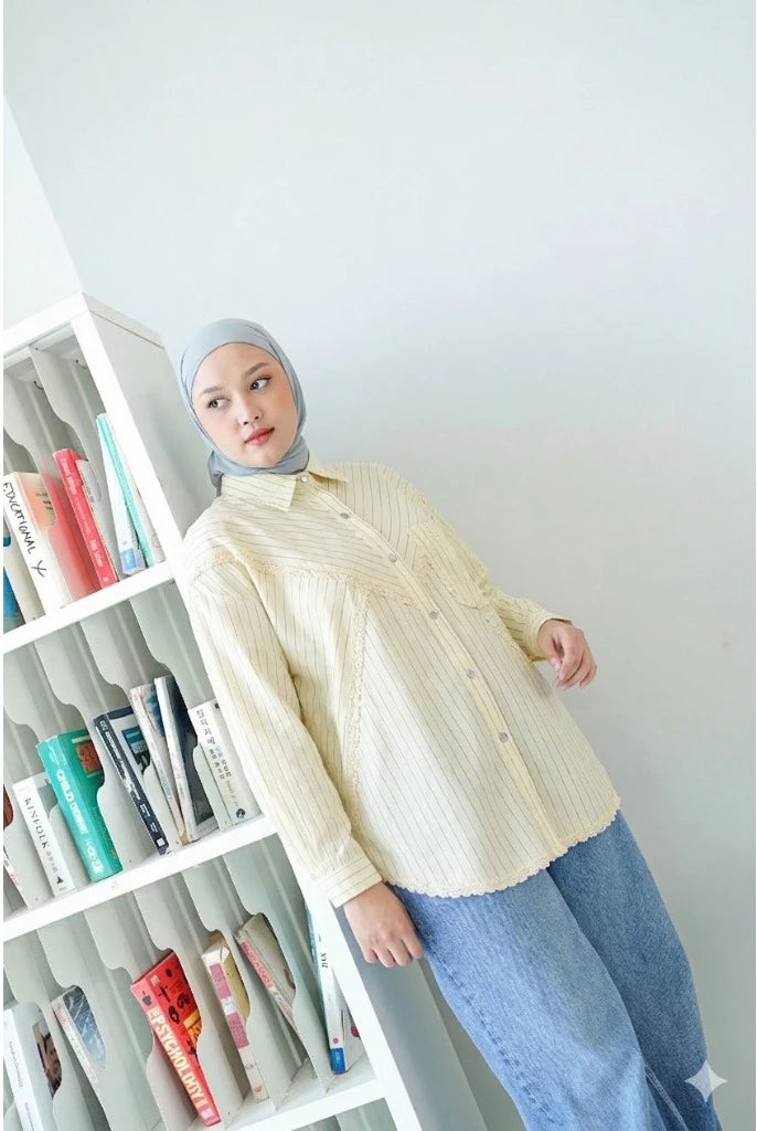 Person wearing a cream shirt by Abame and blue jeans standing in front of a bookshelf.