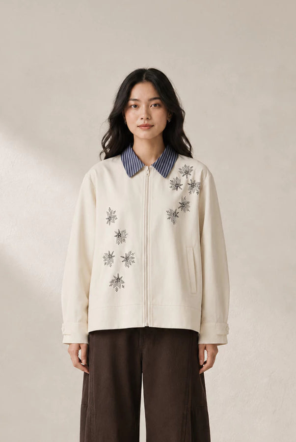 A woman stands against a warm beige wall wearing a cream-colored zip-up jacket by Jenna & Kaia with gray embroidered starburst patterns, a blue-and-white striped collar, and dark brown wide-leg pants.