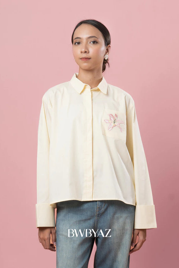 Person wearing a light-colored shirt with small floral embroidery on a pink background, featuring the brand 'BWBYAZ'.