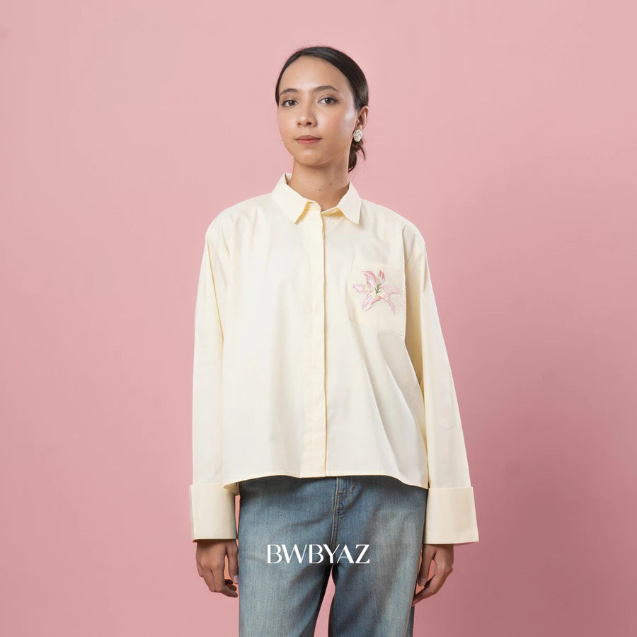 Person wearing a light-colored shirt with small floral embroidery on a pink background, featuring the brand 'BWBYAZ'.