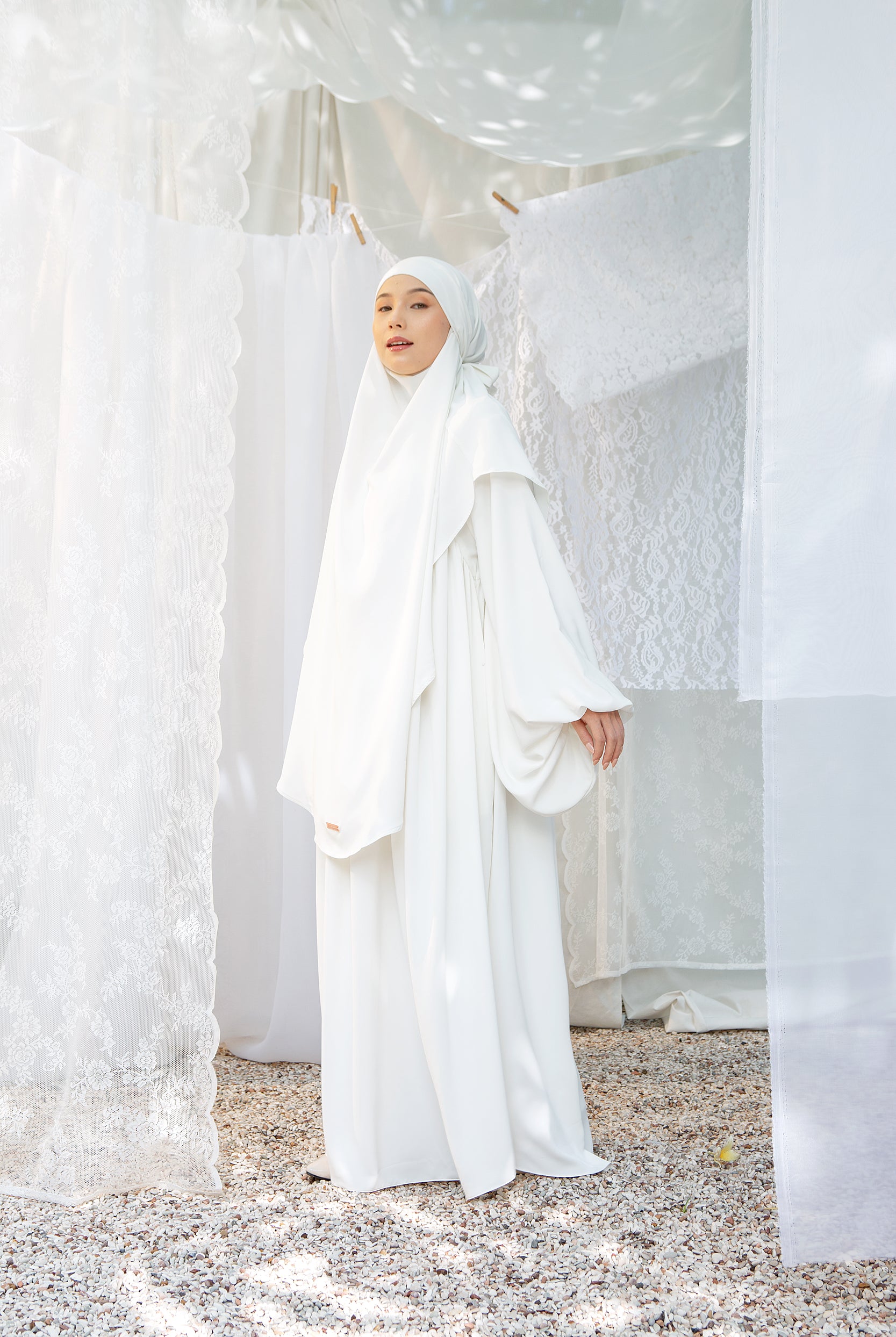 A woman wearing a white abaya set with a matching headscarf by Lozy Hijab, standing indoors.