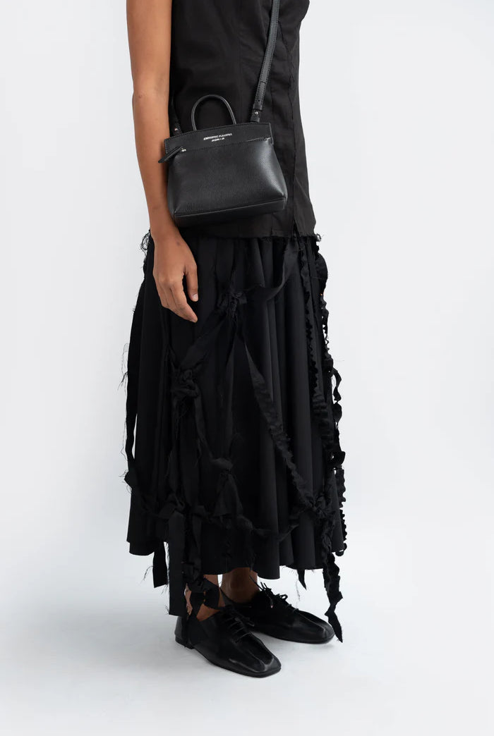 Person wearing a black ruffled skirt and black shoes with a black grain leather handbag by Aesthetic Pleasure on a white background