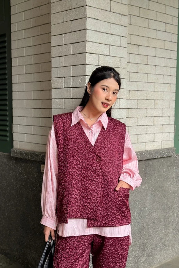 Woman wearing a maroon patterned vest over a pink shirt and pants, standing against a brick wall.