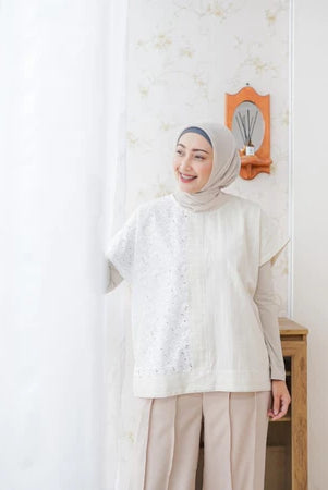 A woman wearing a cream-colored top by Abame with short sleeves and a round neckline, paired with trousers, standing in a room with a light-colored wall and a decorative mirror.