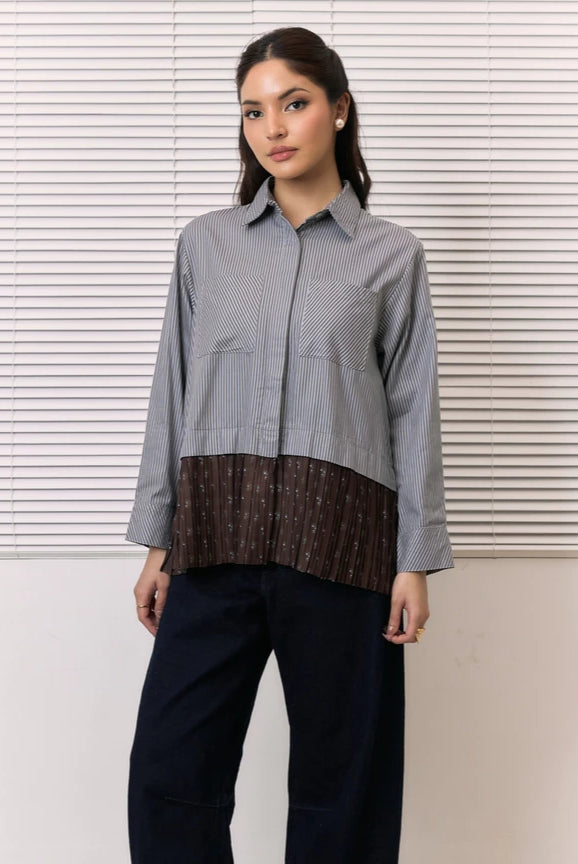 A woman modeling a long-sleeve button-up shirt by Studio Tui with a vertically striped gray and white upper section and a brown pleated lower section with small white floral motifs, paired with dark blue jeans, set against a background of white window blinds.