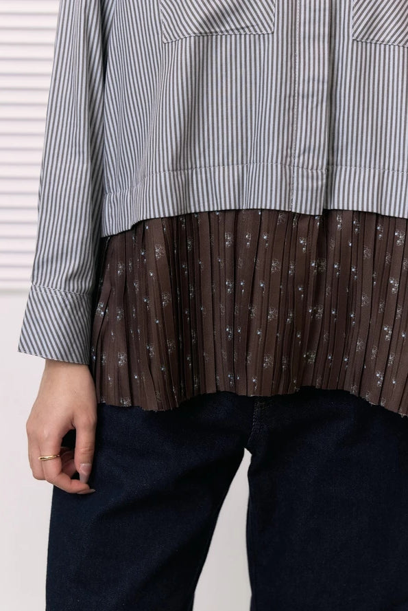 A close-up of a person wearing a long-sleeve shirt by Studio Tui with a vertically striped gray and white upper section and a brown pleated lower section with small white floral motifs, paired with dark blue jeans.